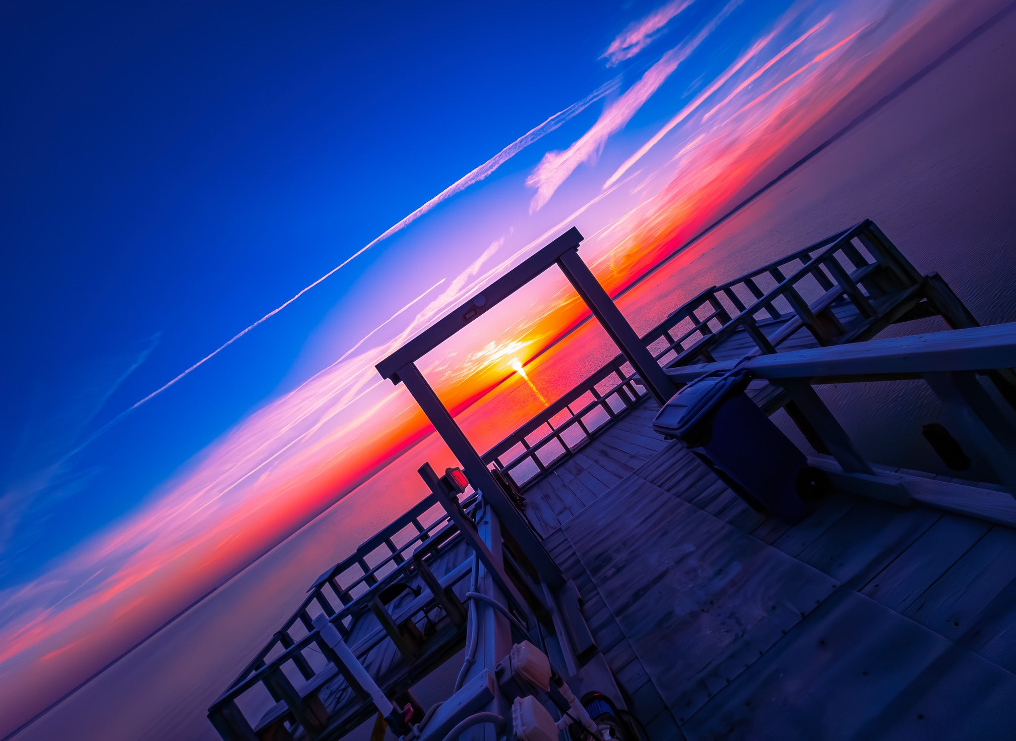 Angled view of a dock at sunrise with a blue and pink sky, wooden railings, and the sun low over calm water.