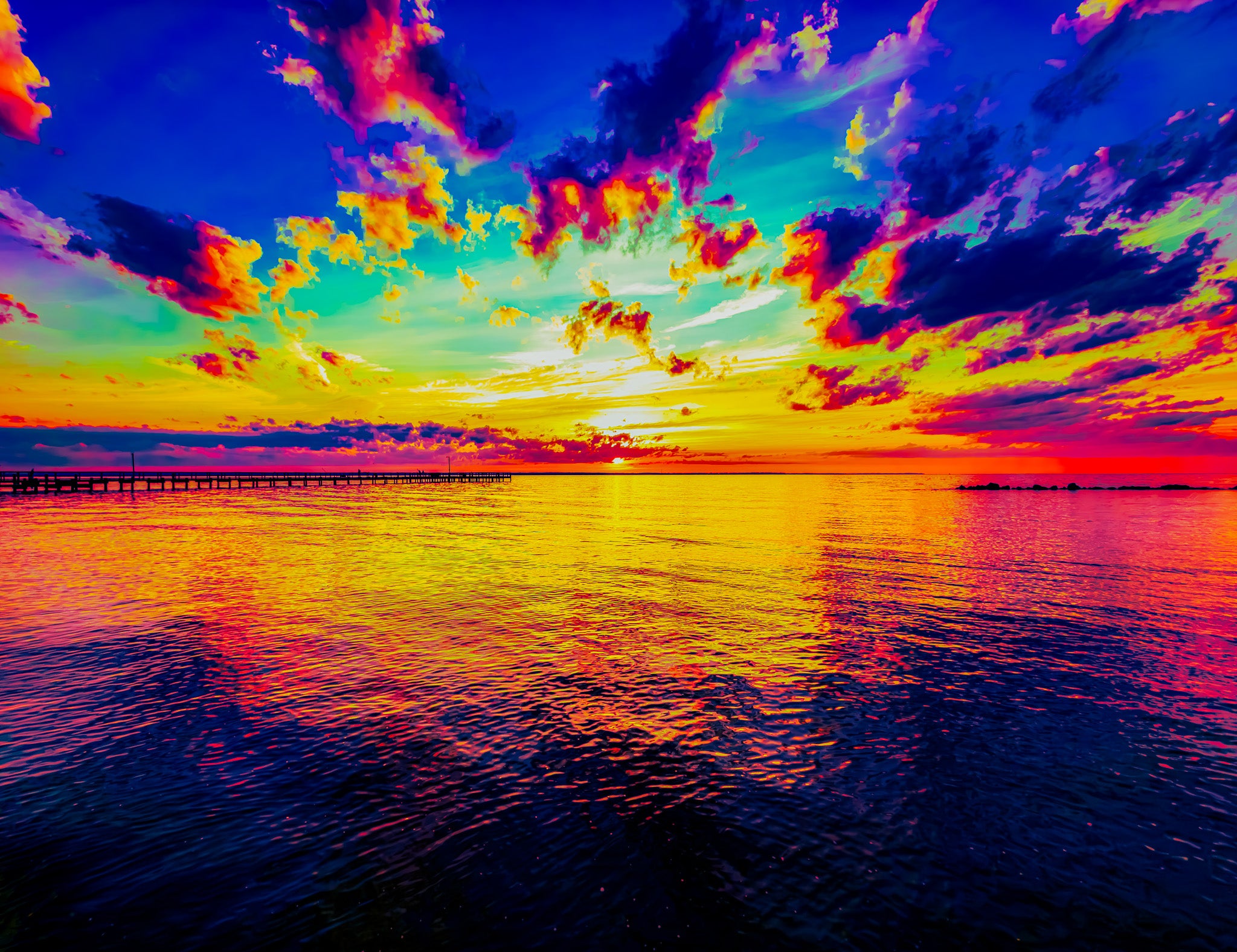 A vivid sunset landscape over calm water, with a long pier extending into the distance. The sky is filled with dramatic clouds in bright blue, purple, pink, orange, and yellow, reflecting strongly across the water surface.