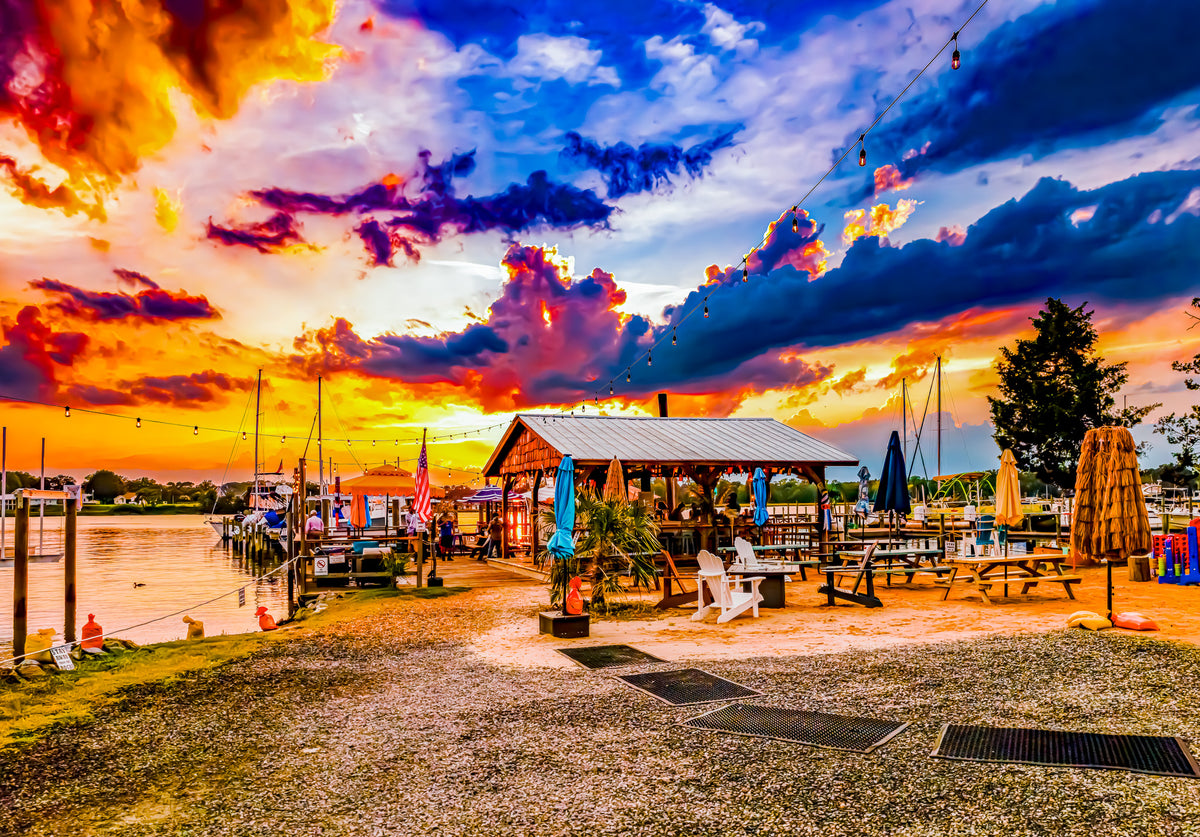 A vibrant outdoor waterfront scene at sunset with a gravel dock path, picnic tables under a pavilion, boats in the water, and a dramatic sky filled with orange, yellow, blue, and purple clouds.
