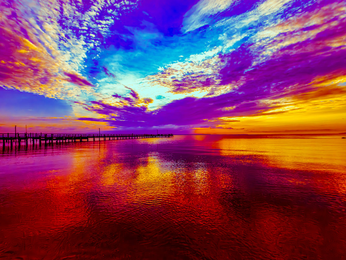 A vivid sunset seascape with a long pier extending into calm water under a dramatic sky filled with purple, blue, orange, and yellow clouds, reflected in the water below.