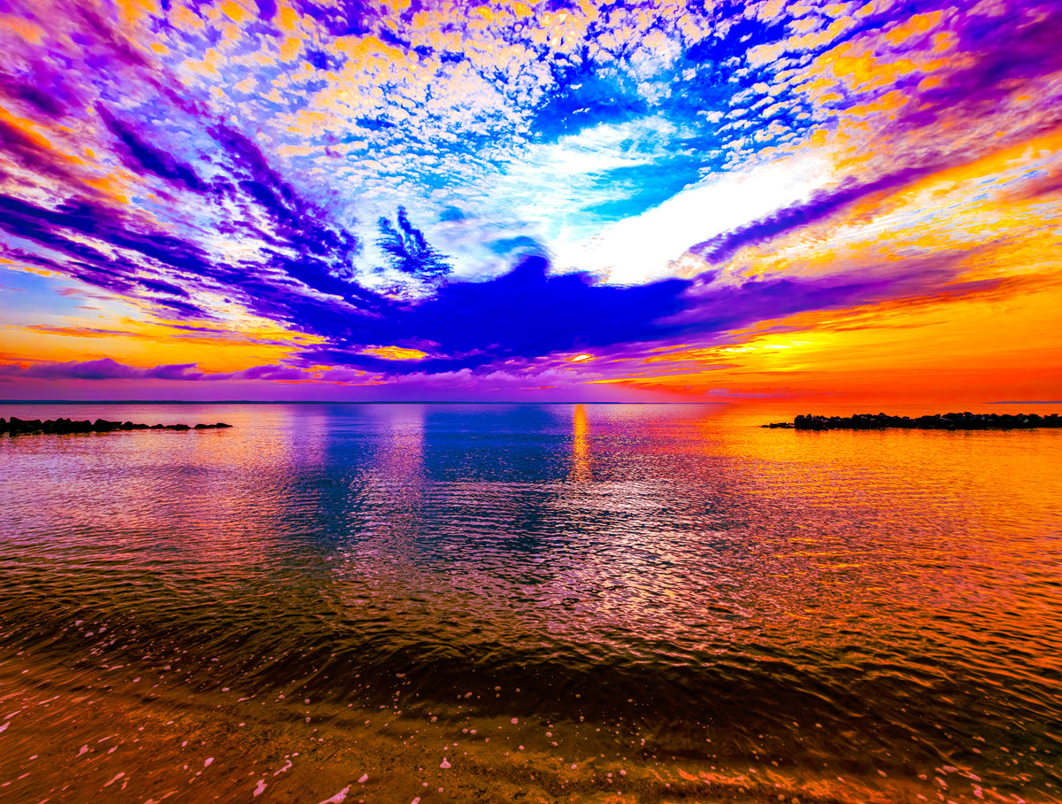 A vivid sunset seascape with a dramatic sky filled with purple, blue, orange, and yellow clouds reflecting over calm water, with dark shoreline silhouettes on both sides.