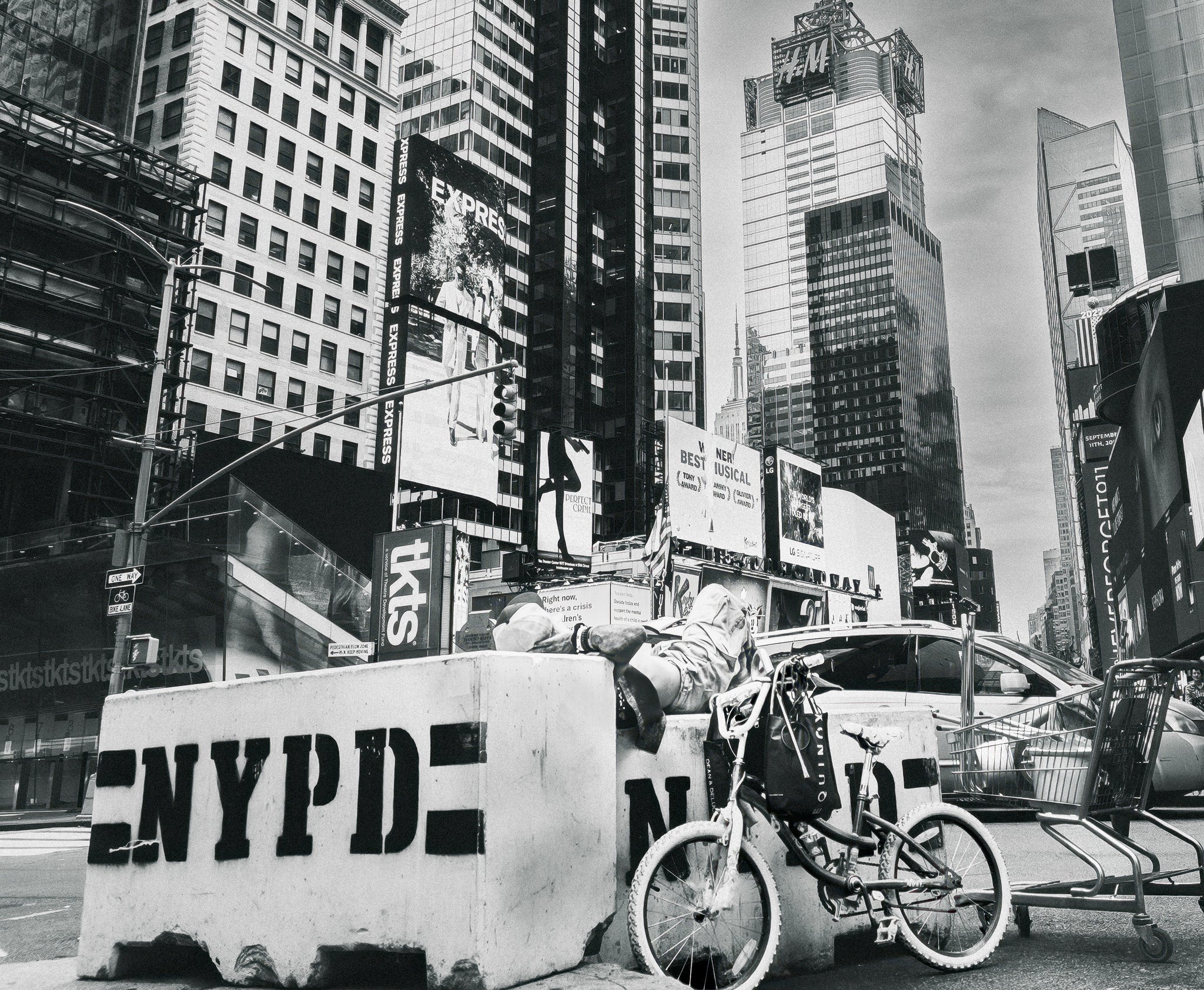 Black-and-white city street scene with man, tall buildings, billboards, an NYPD barrier, a bicycle, and a shopping cart.