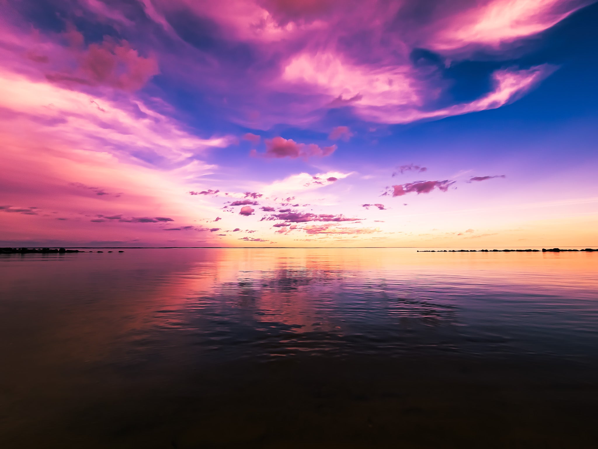 Pink, purple, and blue sky over still water with soft reflections, a low horizon, and rock barriers at the edges.