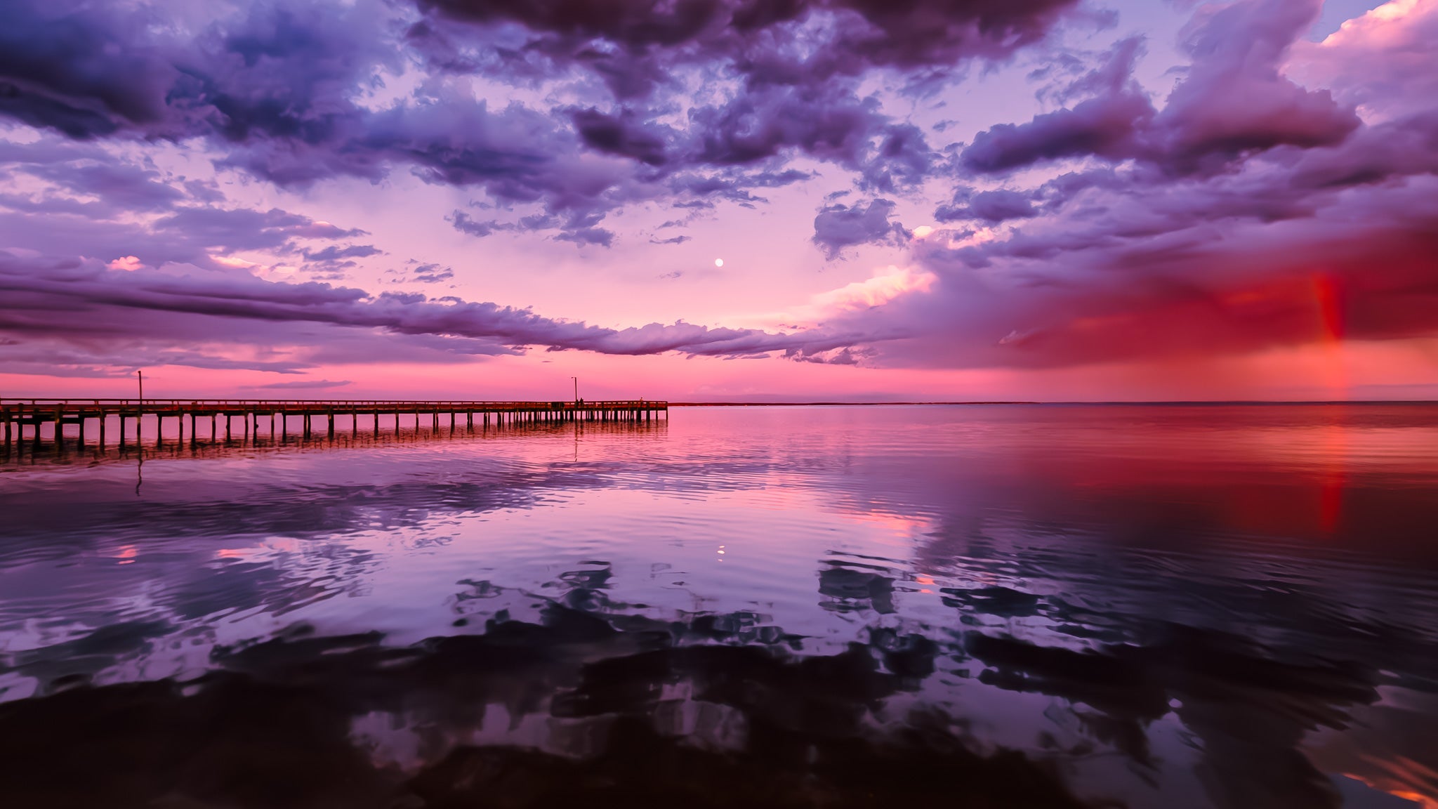 Pink and purple sky over still water with cloud reflections, a long pier on the left, and a faint rainbow on the right.