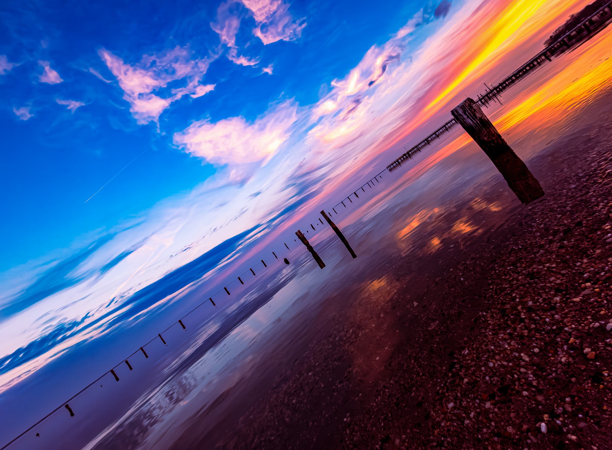 Angled view of a blue and pink sky over shallow water with scattered pilings, a pebbled shoreline, and a pier near the horizon.