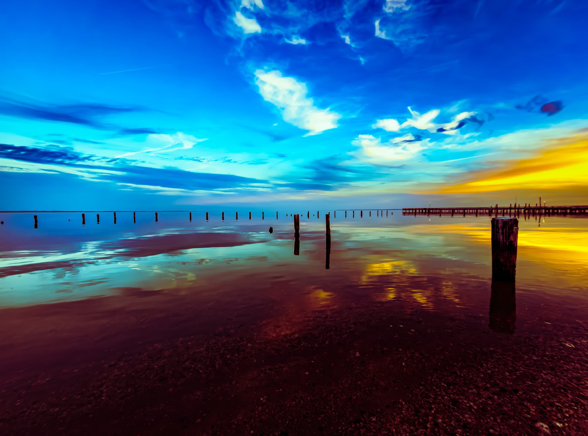 Blue sky over still water with reflections, scattered pilings, and a pier on the right near a bright yellow horizon.