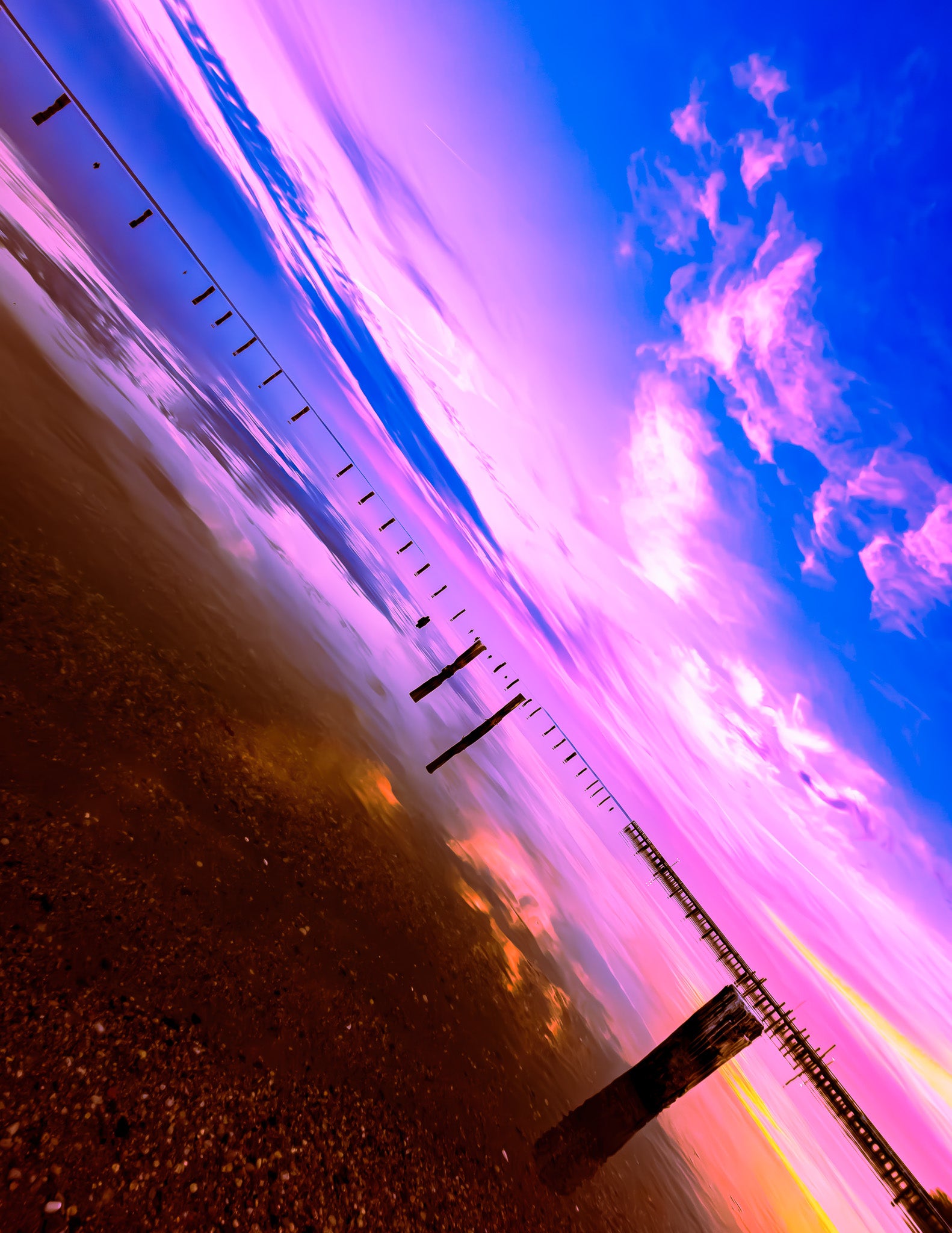 Pink and blue sky over shallow water with reflections, scattered pilings, and a pier on the right.
