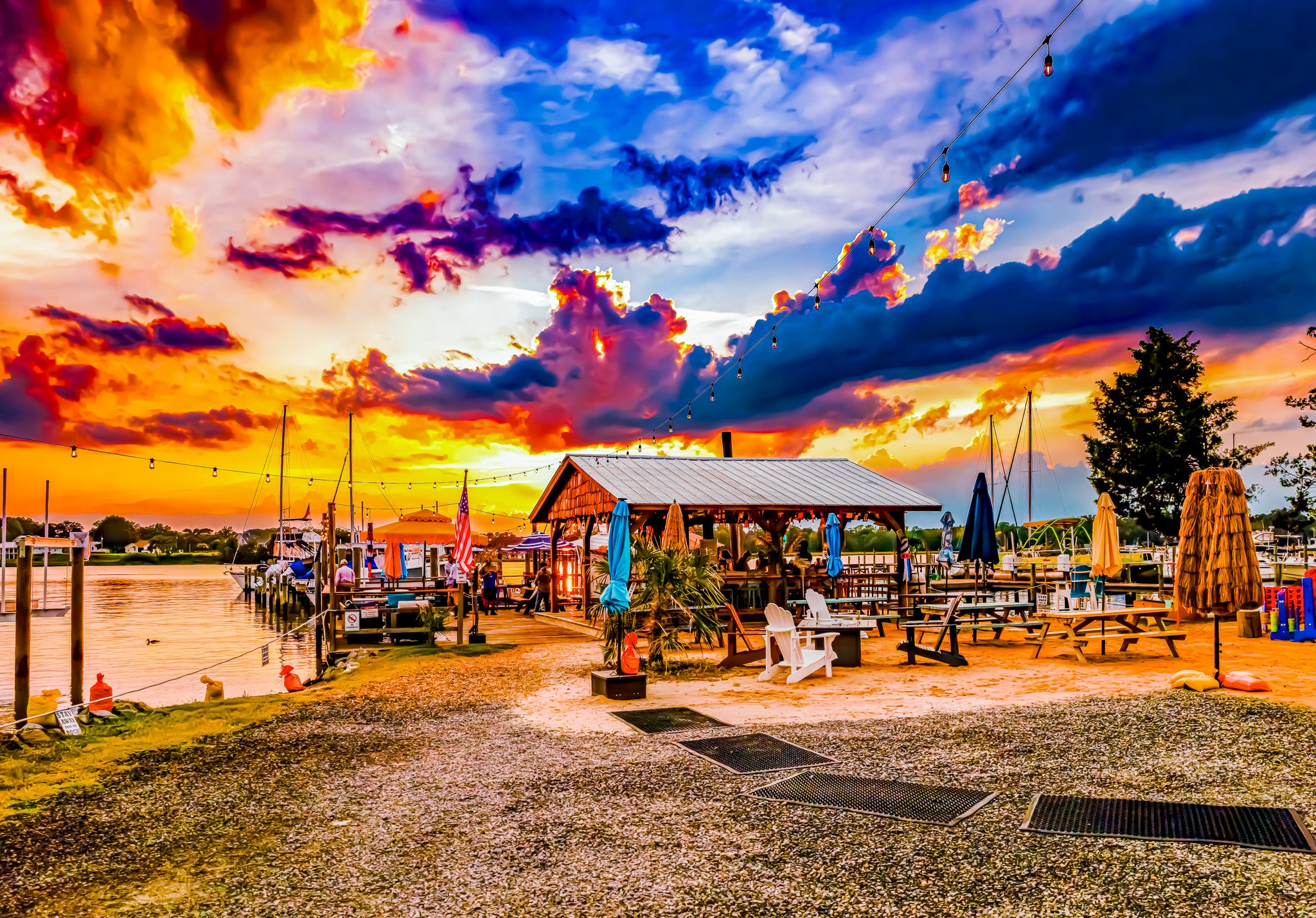 A vibrant outdoor waterfront scene at sunset with a gravel dock path, picnic tables under a pavilion, boats in the water, and a dramatic sky filled with orange, yellow, blue, and purple clouds.