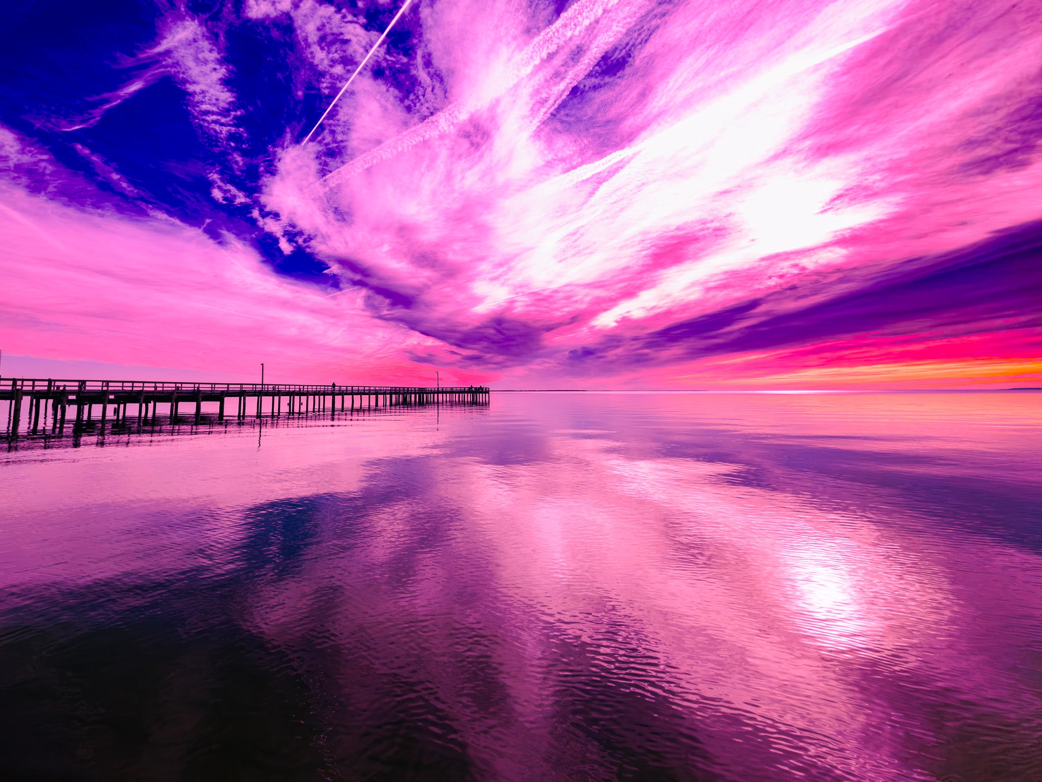 Colorful sunset over a pier with reflection on water. Untitled — The Sky Isn’t Falling…Yet 008 by Austin Red. Pink and purple sky-and-water photography with reflection and a long pier, produced with WhiteWall using gallery-grade materials.”